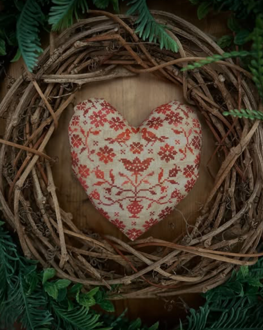 A heart-shaped decorative pillow with a red floral pattern on a cream fabric background, nestled inside a rustic twig wreath with green foliage.