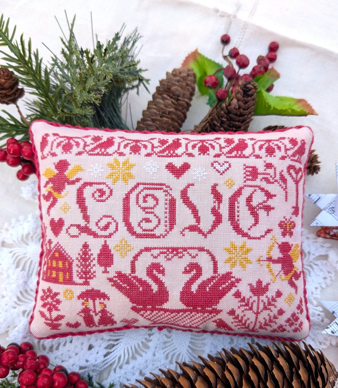 Decorative pillow with cross-stitch design featuring swans and the word 'love', surrounded by Christmas decorations.