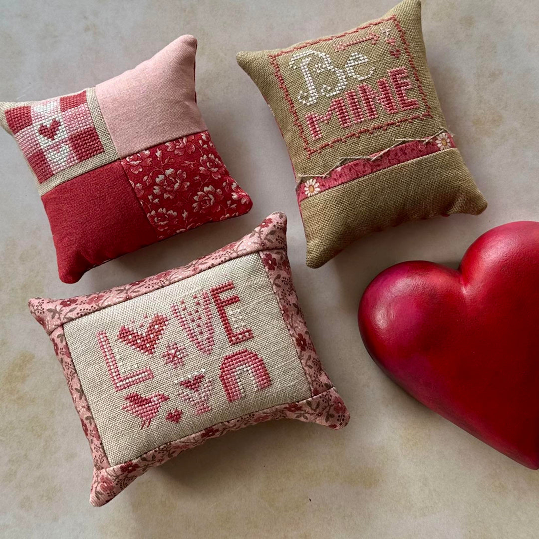 Three decorative pillows with embroidered text and a red heart on a marble surface
