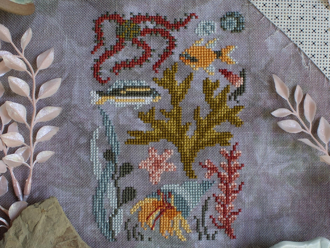 Cross-stitched design of underwater scene with fish and plants on gray fabric.