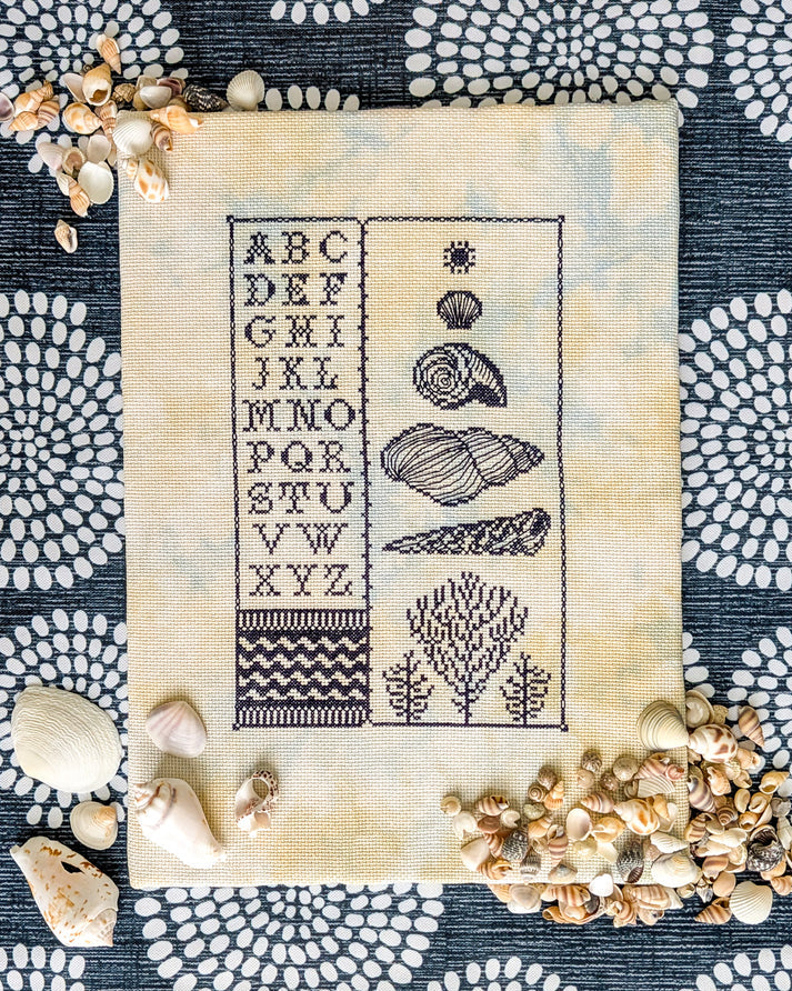 A square cream fabric sampler or cross-stitch piece with a navy alphabet and seashell motif, laid flat on a dark patterned surface and surrounded by real seashells.