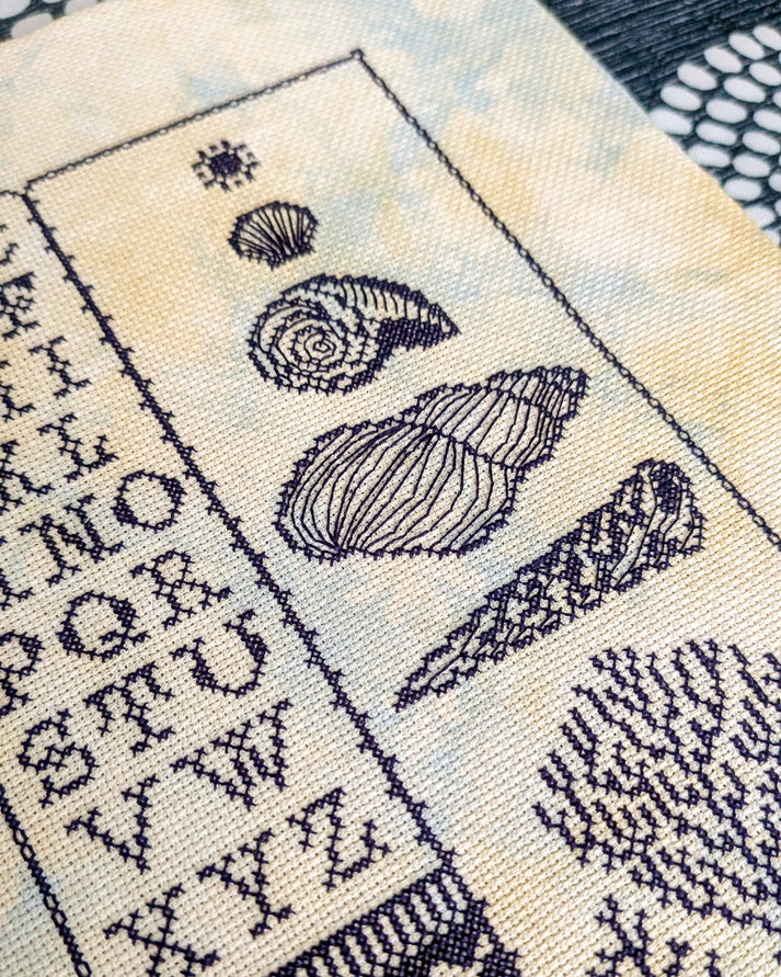 Close-up of a needlepoint canvas featuring a navy blue alphabet and shell motifs on a cream background with a subtle yellow and blue mottled texture.