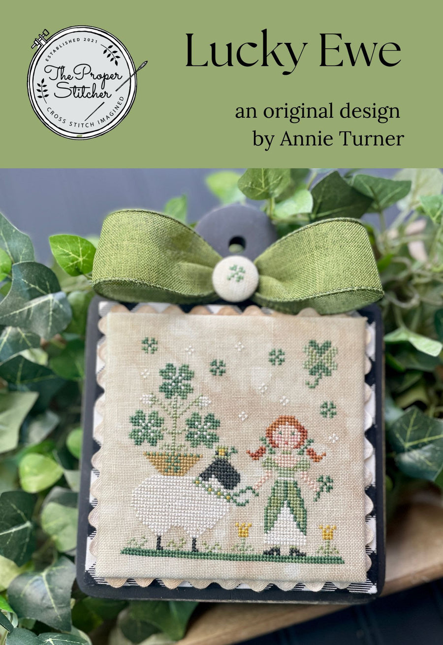 Cross-stitched design of a shepherd and sheep with greenery, branded 'Lucky Ewe' by Annie Turner.