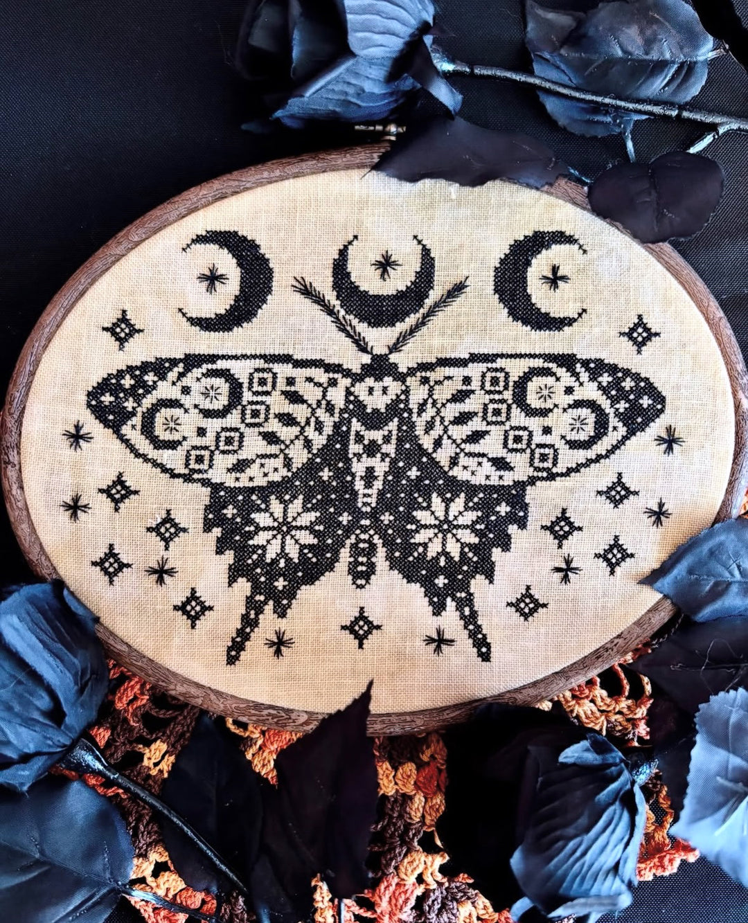 A circular embroidery piece on a wood hoop featuring a black cross-stitched moth with patterned wings, three crescent moons, and small star-like motifs on a beige fabric background, styled with dark blue feathers and dried botanicals.