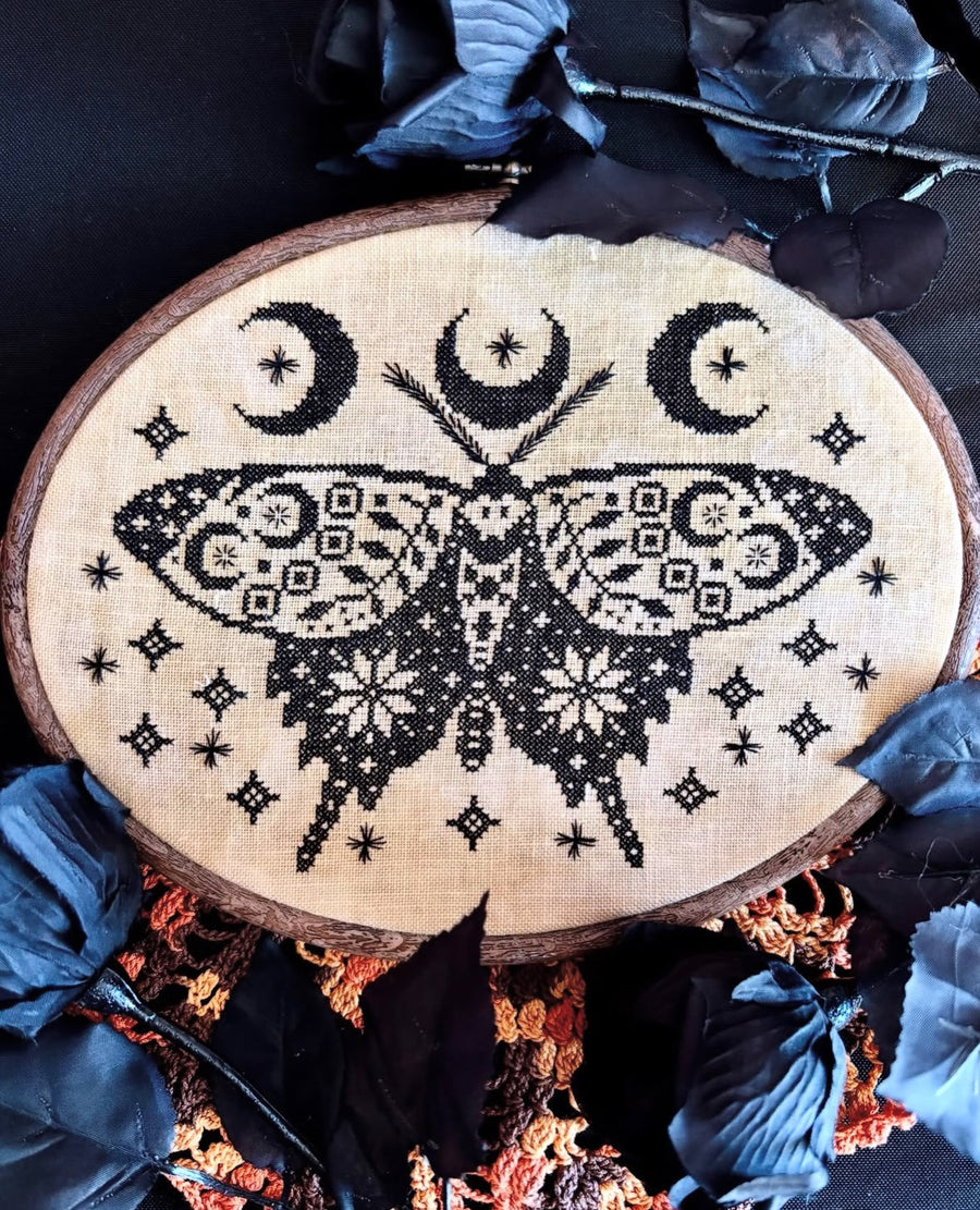 A circular embroidery piece on a wood hoop featuring a black cross-stitched moth with patterned wings, three crescent moons, and small star-like motifs on a beige fabric background, styled with dark blue feathers and dried botanicals.