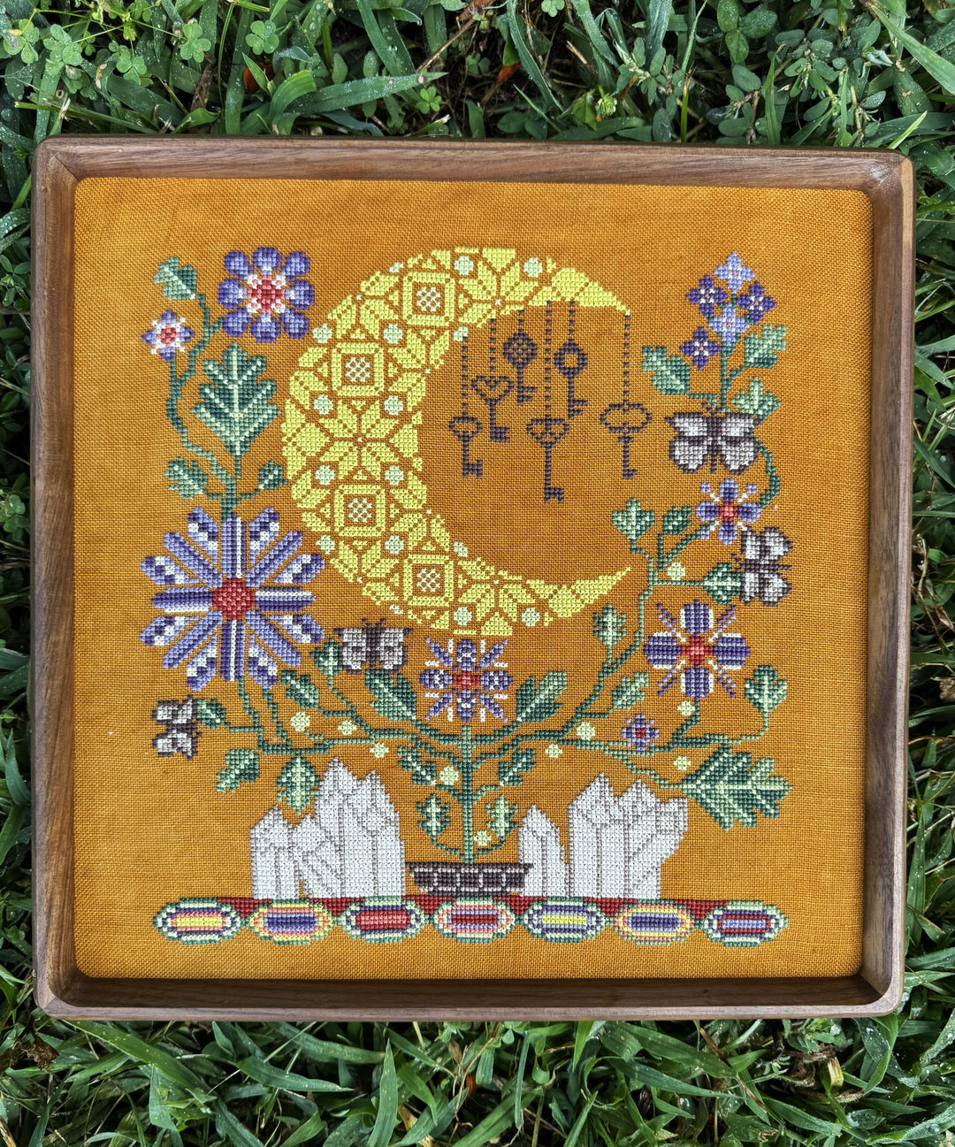A framed cross-stitch artwork featuring a yellow crescent moon, hanging keys, flowers, and birds on a mustard-yellow fabric background, placed outdoors on green grass.