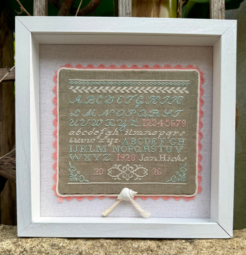 A framed vintage-style cross-stitch sampler featuring an alphabet, numbers, and decorative borders. The sampler is mounted on a beige fabric background with a pink scalloped edge, set inside a white wooden frame.