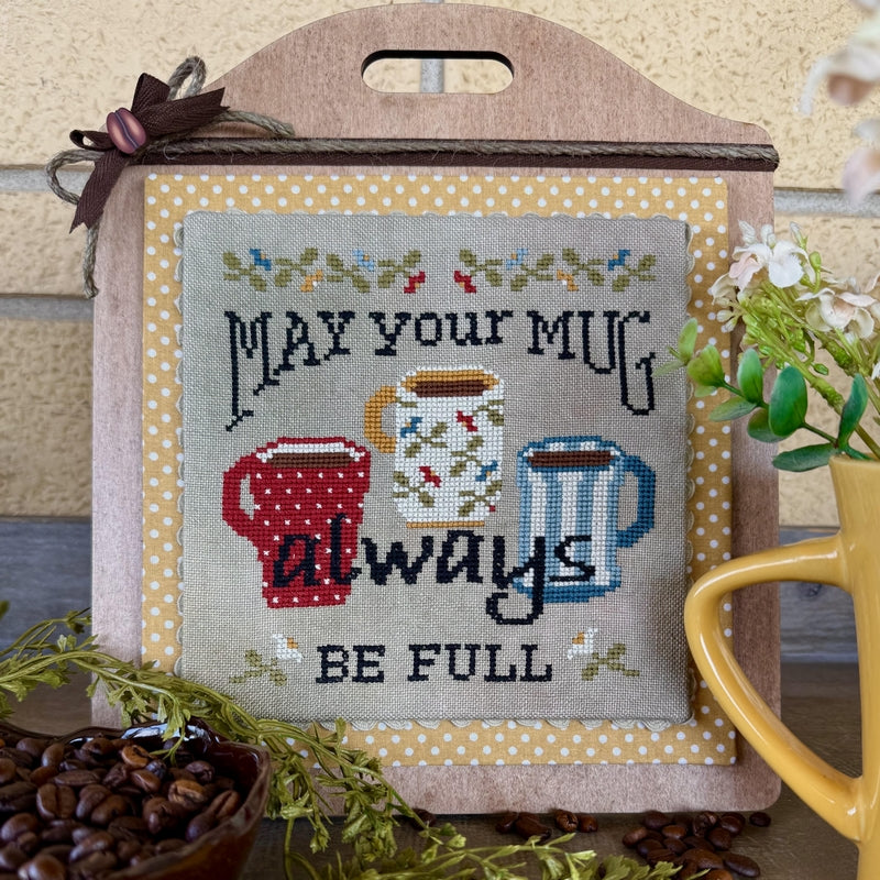 A decorative cross-stitch sampler featuring the phrase 'MAY YOUR MUG always BE FULL' with three illustrated mugs, displayed on a wooden stand with a ribbon and surrounded by coffee beans, greenery, and a yellow mug.