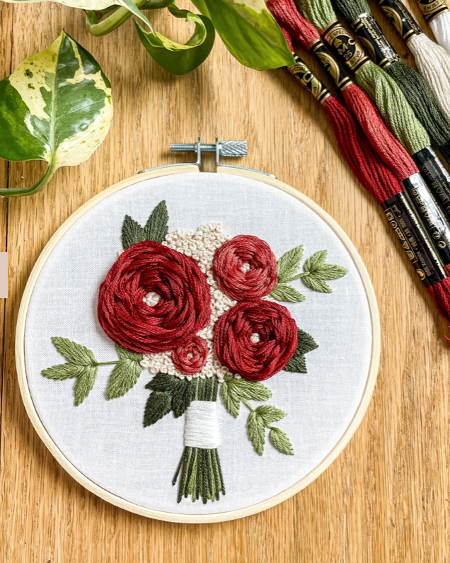 A round embroidery hoop displays a bouquet of red and burgundy roses with green leaves stitched on white fabric, set on a wooden surface with embroidery floss skeins and green leaves in the background.