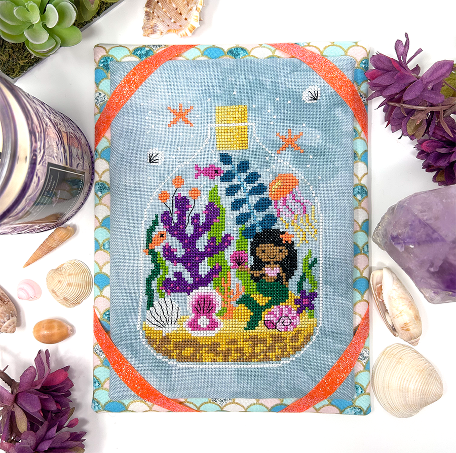 Cross-stitched mermaid in an underwater scene with seashells and flowers on a white background