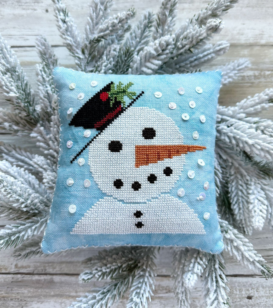 Cross-stitched snowman pillow on a wooden surface with pine branches