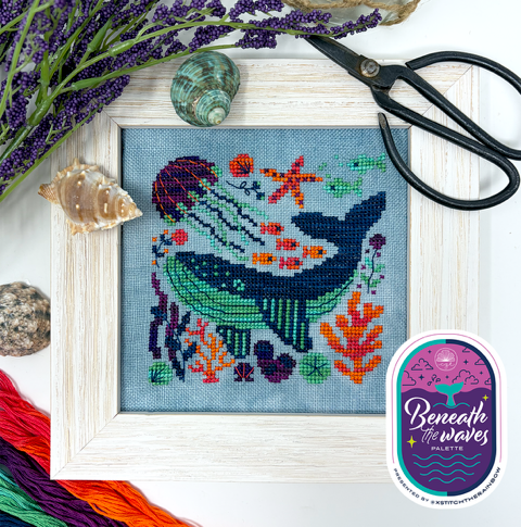 Cross-stitched whale design with embroidery floss, scissors, and 'Beneath the Waves' palette on a white background.
