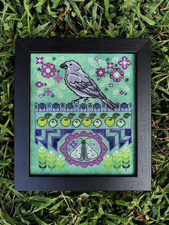 A framed cross-stitch artwork featuring a bird, flowers, and decorative patterns on a mint green background, placed on grass.