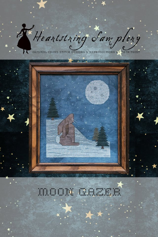 Heartstring Samplery 'Moon Gazer' cross stitch pattern cover, featuring a framed winter night scene with a full moon, pine trees, and a house on a snowy ground, set against a dark starry sky.