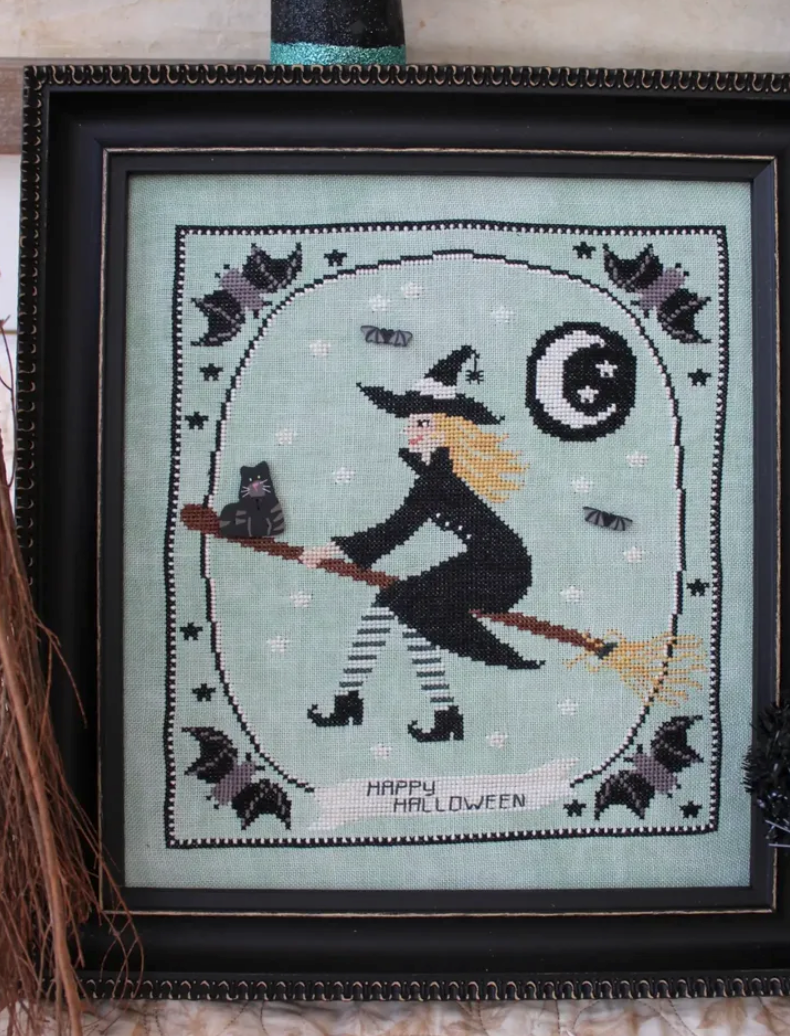 A framed needlepoint or cross-stitch artwork featuring a witch riding a broomstick with a cat, stars, bats, and a crescent moon on a pale mint background, with the words “HAPPY HALLOWEEN” stitched at the bottom. The piece is enclosed in a black frame.