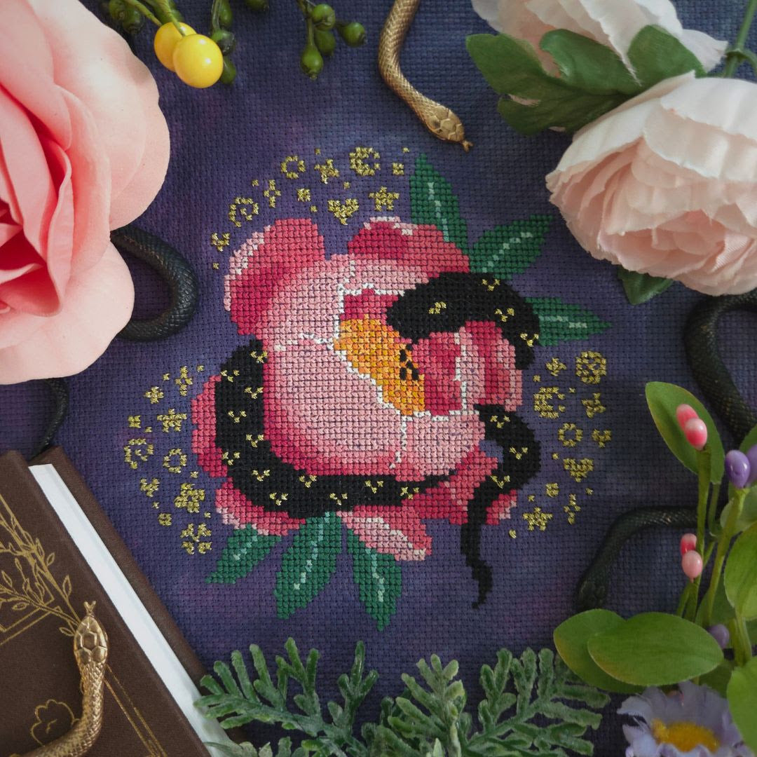 A close-up of a counted cross-stitch pattern depicting a pink rose with a black snake on a dark purple fabric background, surrounded by decorative pink flowers, greenery, and a book corner.