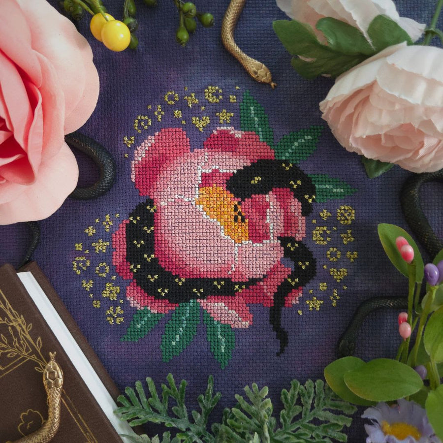 A close-up of a counted cross-stitch pattern depicting a pink rose with a black snake on a dark purple fabric background, surrounded by decorative pink flowers, greenery, and a book corner.