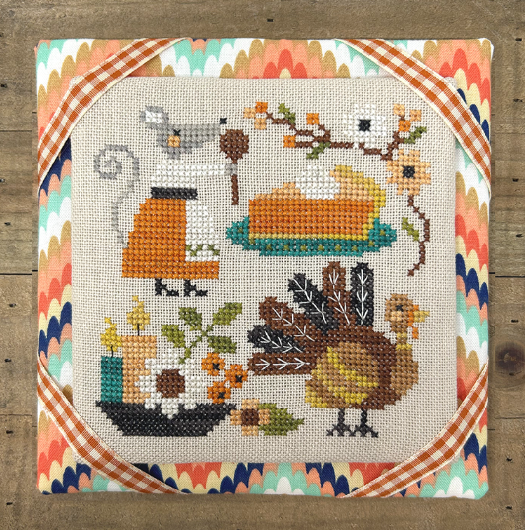 Cross-stitched piece with a turkey, pie, and other fall-themed elements on a wooden frame.