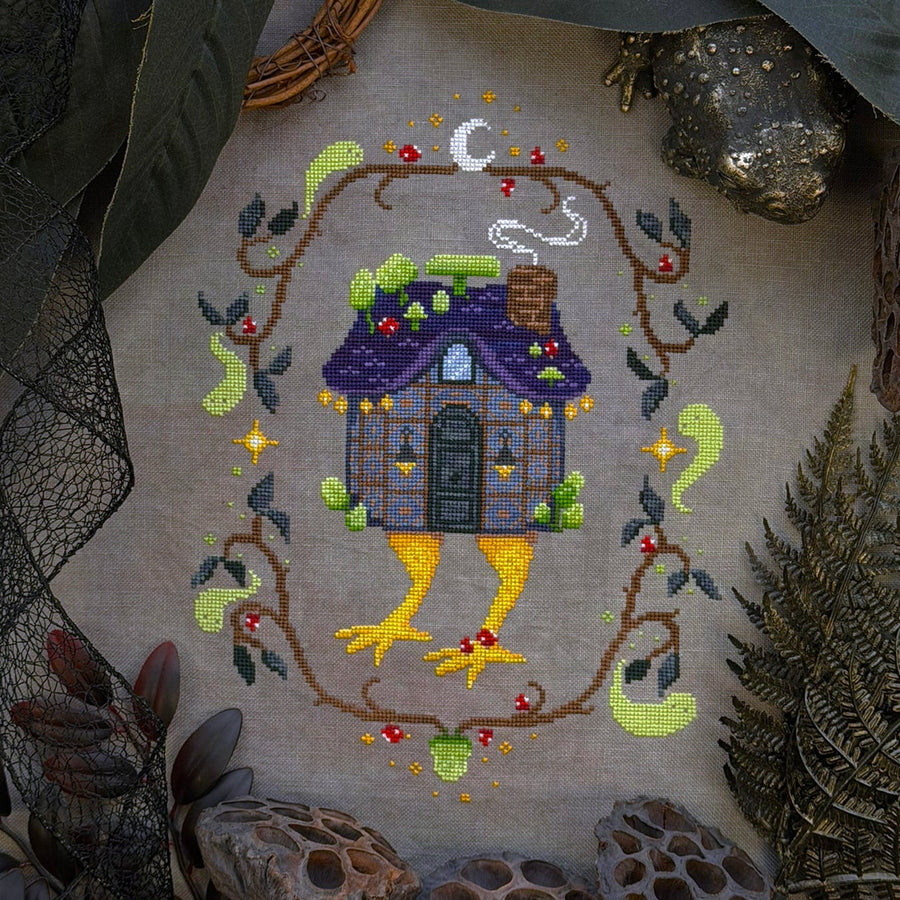 A cross-stitch or needlework design of a whimsical fairy house with yellow legs, framed by a leafy vine border, displayed on a textured fabric surrounded by natural props like leaves, moss, and dried flowers.