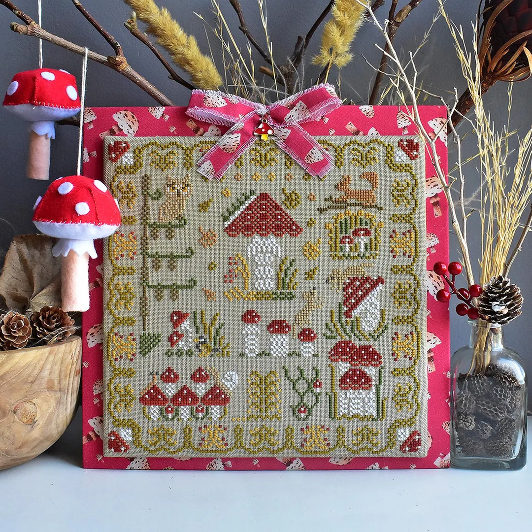 Cross-stitched pattern with mushroom design on a pink border, surrounded by decorative elements.