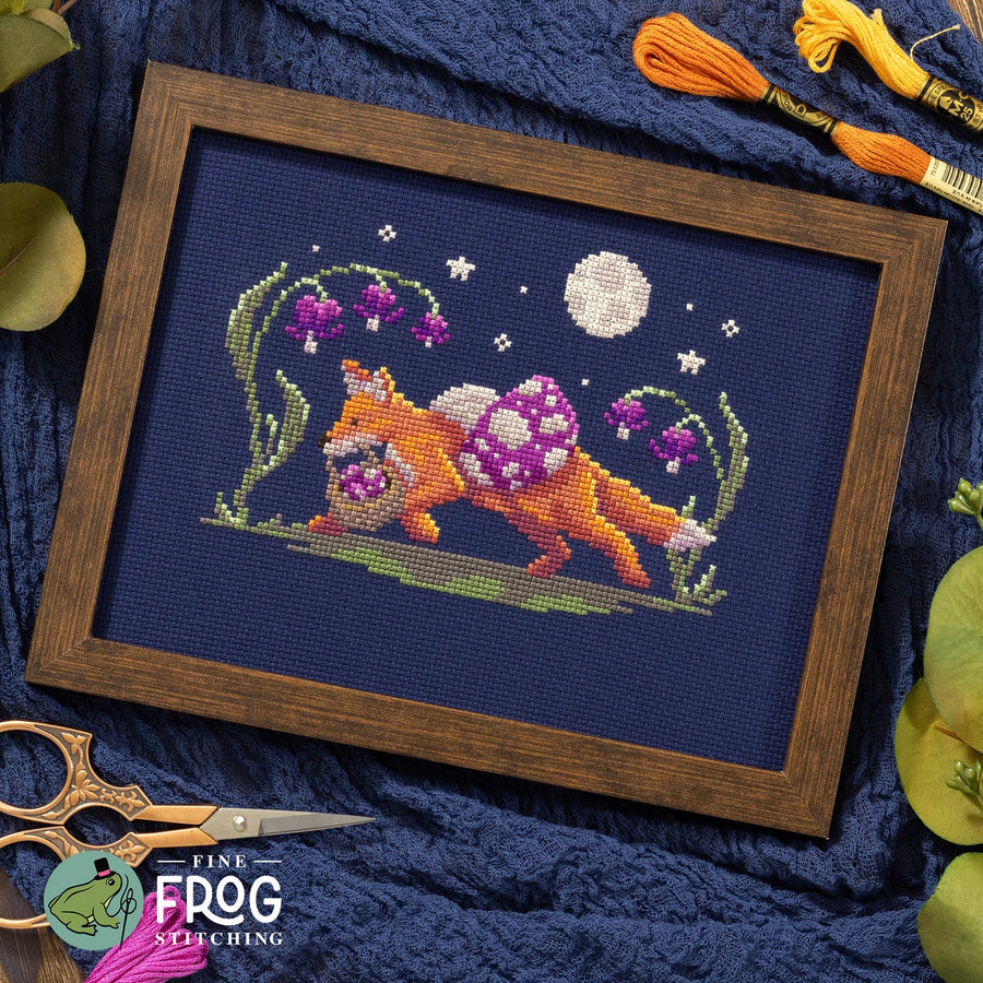 A framed cross-stitch artwork showing an orange fox carrying a purple-and-white spotted sack on its back, surrounded by purple flowers and a white moon on a dark blue background. The piece is displayed in a brown wooden frame on a textured navy fabric, with tinsel, scissors, and thread visible around it.