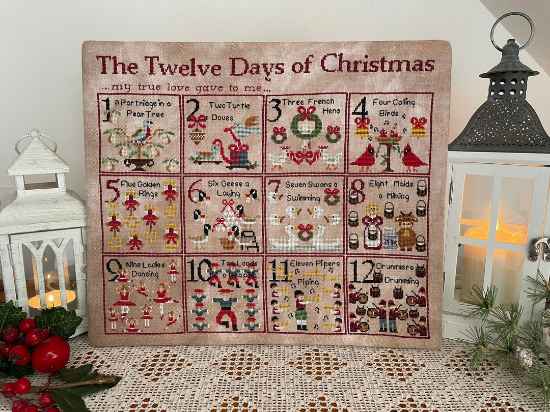 A framed or mounted fabric Advent calendar titled "The Twelve Days of Christmas" displayed on a lace-covered surface, surrounded by decorative lanterns, candles, and holiday greenery.