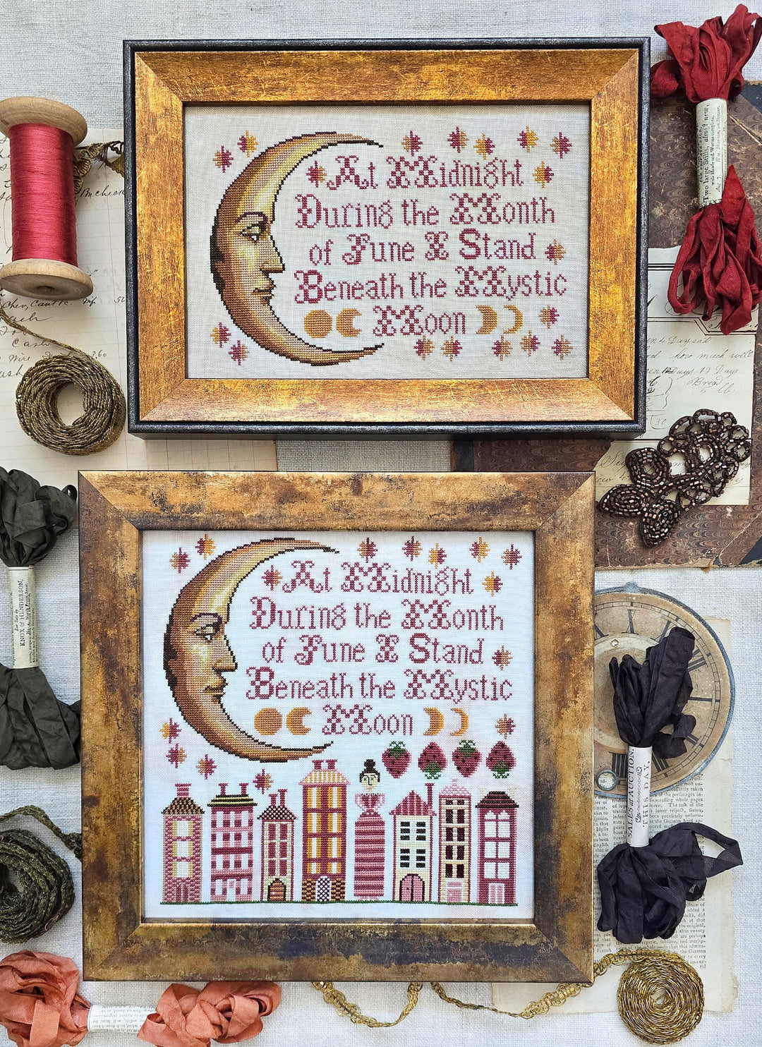 Two framed cross-stitched pieces with moon and cityscape designs, surrounded by decorative ribbons and threads.