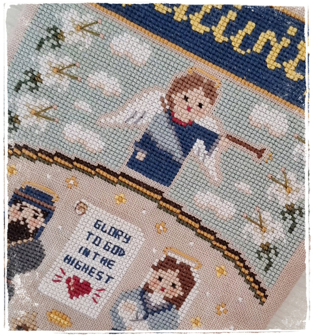 A close-up of a finished cross-stitch needlework piece featuring angels, clouds, stars, and the text 'GLORY TO GOD IN THE HIGHEST.' The fabric shows a light blue-gray background with stitched figures in blue, beige, brown, red, and gold tones.