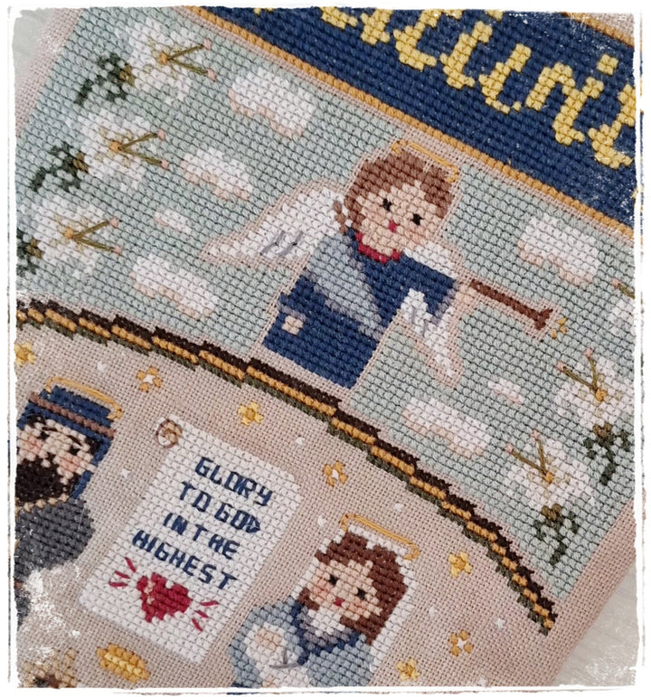 A close-up of a finished cross-stitch needlework piece featuring angels, clouds, stars, and the text 'GLORY TO GOD IN THE HIGHEST.' The fabric shows a light blue-gray background with stitched figures in blue, beige, brown, red, and gold tones.