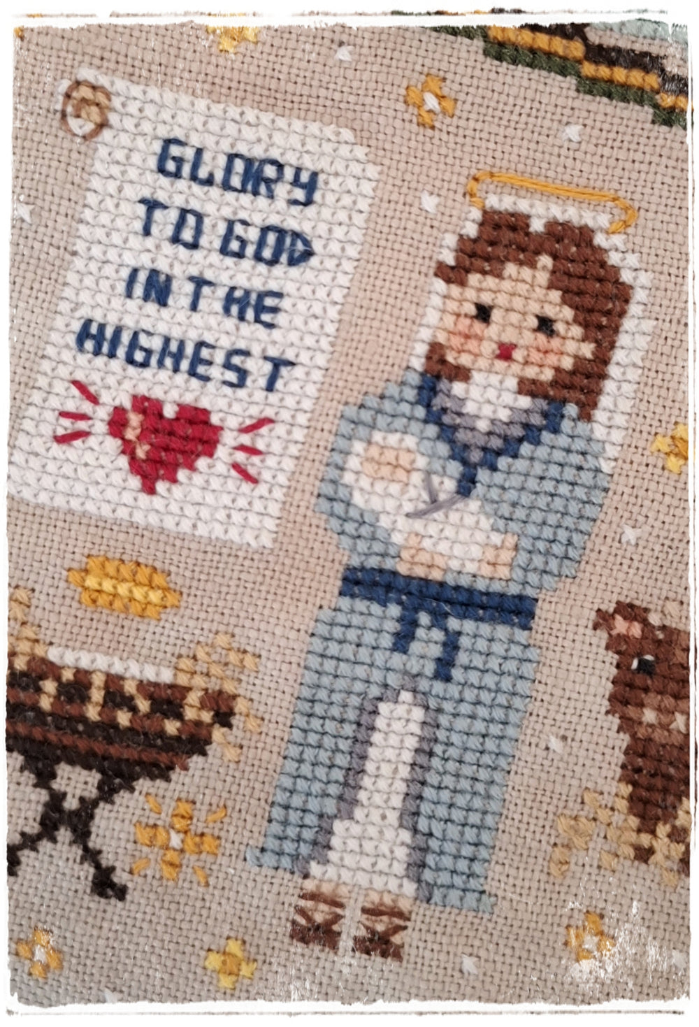 A cross-stitch artwork featuring a nurse in light blue with a stethoscope, holding a sign that reads 'GLORY TO GOD IN THE HIGHEST' with a red heart. The design is on a beige fabric background with small gold stars and brown animal silhouettes.
