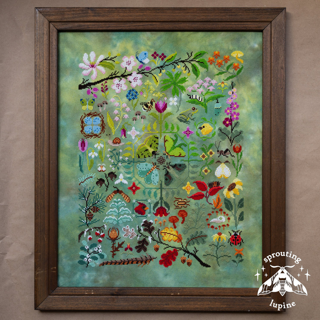 A framed floral needlepoint or embroidery artwork featuring a dense arrangement of wildflowers, leaves, birds, and butterflies on a textured green background, enclosed in a dark brown wooden frame.