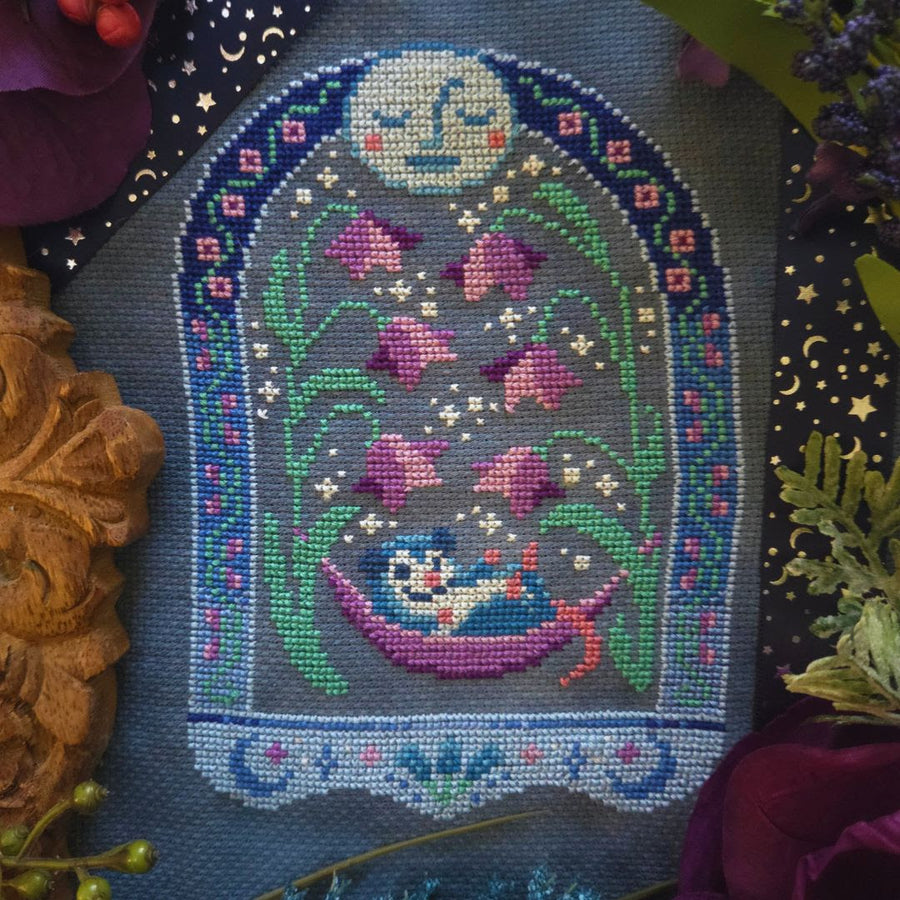 A close-up of a finished cross-stitch or needlepoint textile featuring a whimsical moon face, pink flowers, green vines, and a blue-and-orange bird in a hammock, surrounded by decorative floral and carved props.