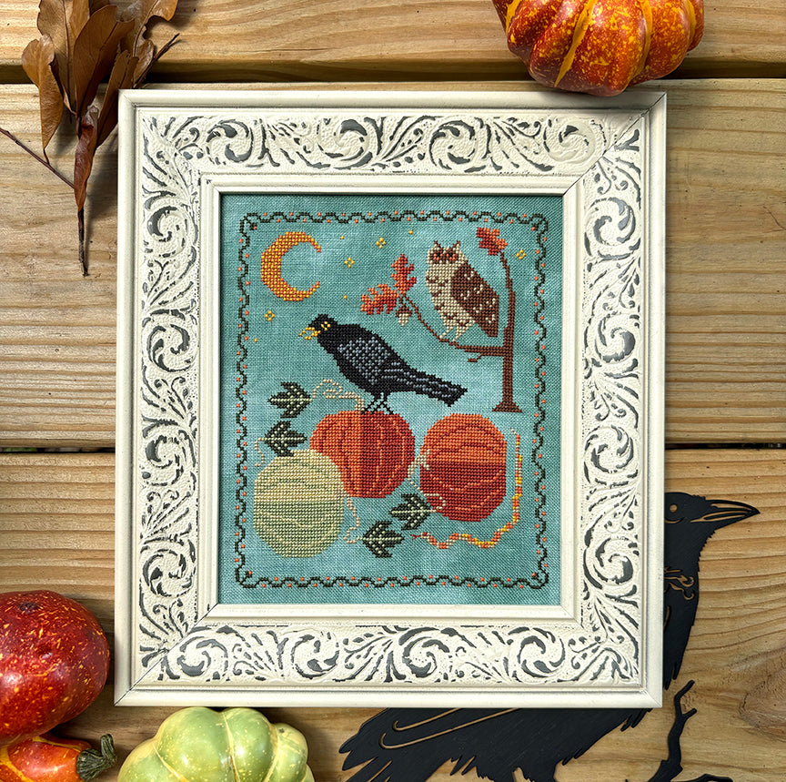 Decorative cross-stitch piece with pumpkins, an owl, and a crow on a wooden surface.
