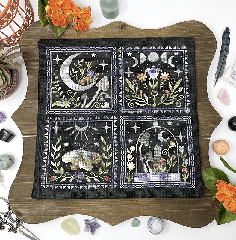Embroidery hoop with cross-stitched design featuring moon, flowers, and animals on a wooden surface.