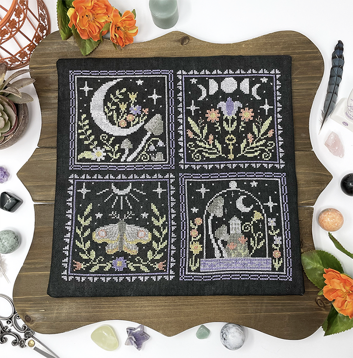 Embroidery hoop with cross-stitched design featuring moon, flowers, and animals on a wooden surface.