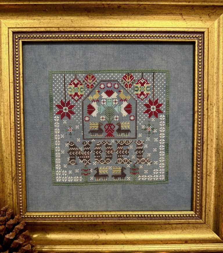 A framed needlepoint artwork featuring a house with birds, floral motifs, and the word "NOR" stitched at the bottom, set in a blue-gray fabric and enclosed in an ornate gold frame.