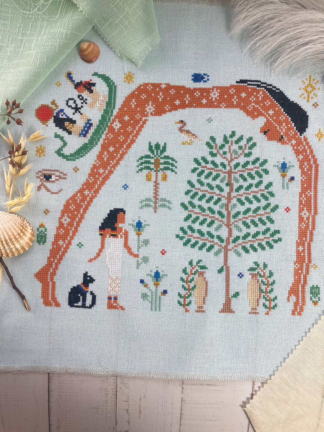 Cross-stitched design of a mermaid, tree, and other elements on fabric.