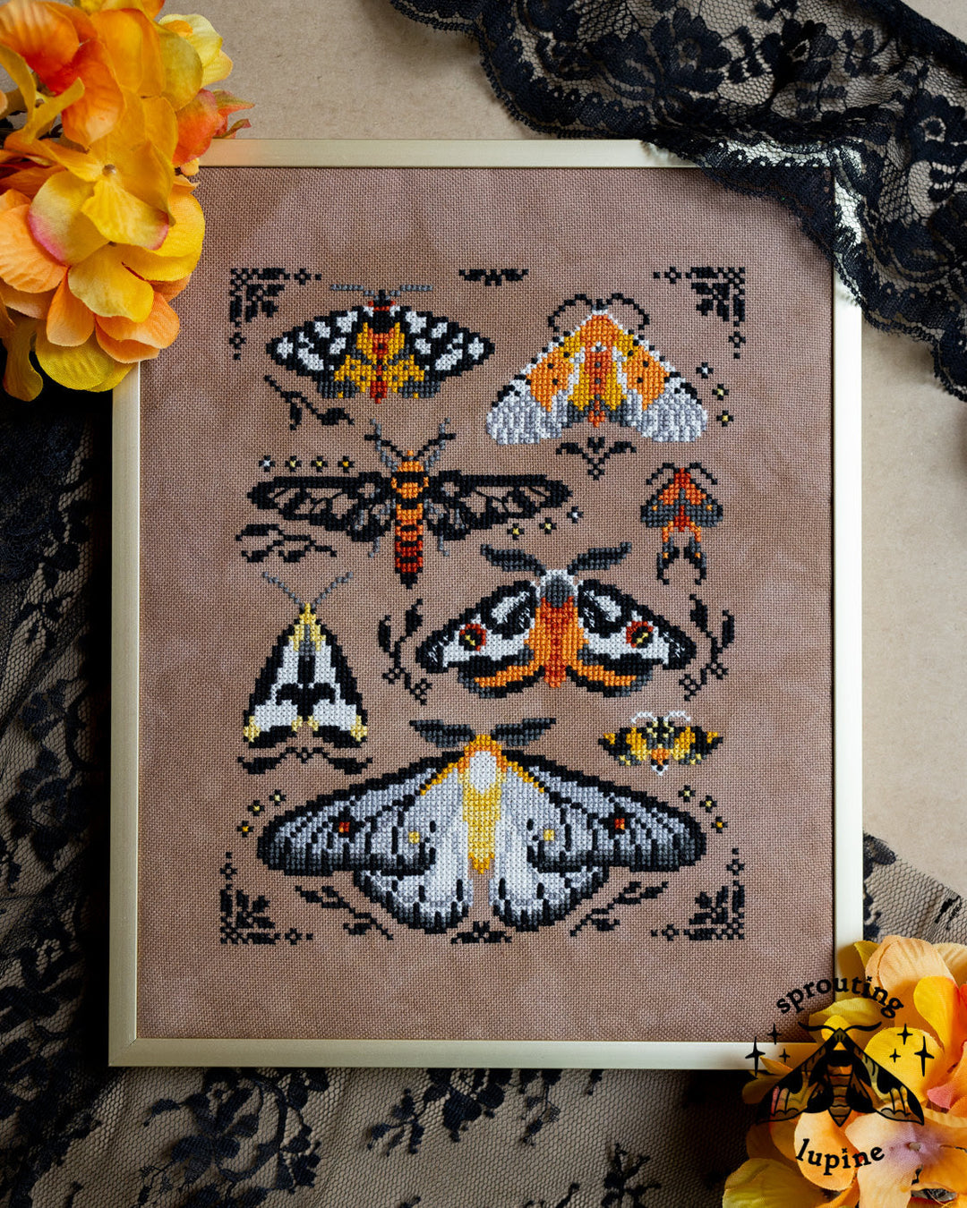 A framed cross-stitch artwork featuring multiple moths and butterflies stitched on a taupe fabric background, styled with black lace and orange flowers around it.