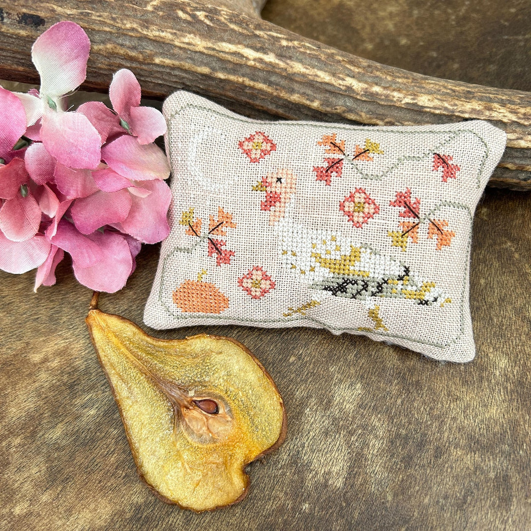 A small rectangular cross-stitch pillow with a floral and bird design, styled on a rustic wooden surface with pink flowers and a dried pear slice.