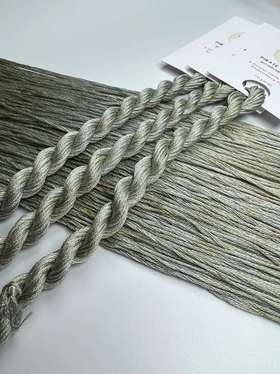 Three skeins of gray green embroidery floss on a white surface with a brand label in the background.