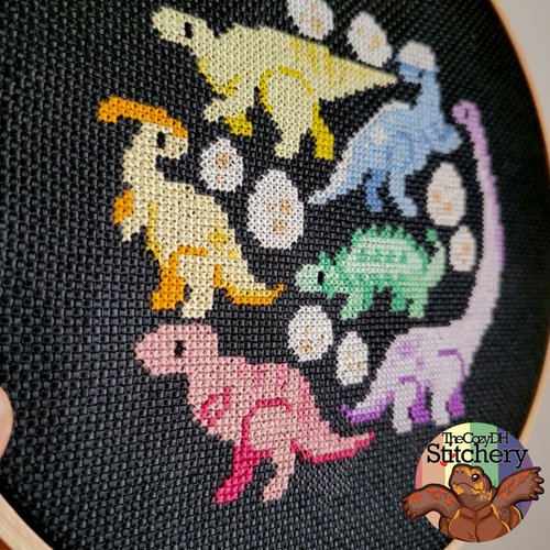 A close-up of a circular black cross-stitch piece featuring colorful embroidered dinosaurs in yellow, blue, green, pink, and purple, with white eggs on a black fabric background. A logo for 'The Crafty Stitchery' is visible in the bottom right corner.