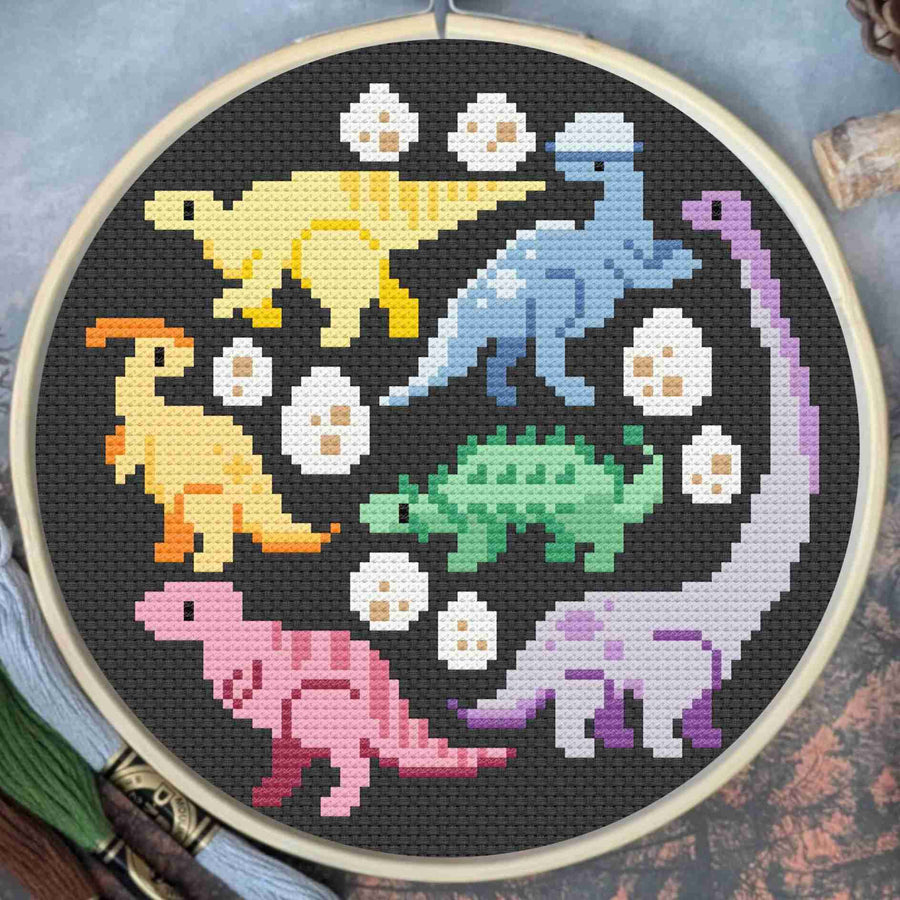 A circular embroidery hoop containing a colorful cross-stitch design of six pixelated dinosaurs and white eggs on a black fabric background, photographed flat with a blurred rustic surface around it.