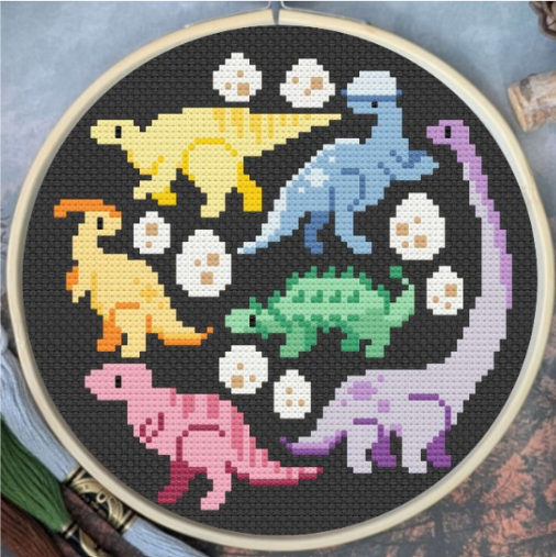A circular embroidery hoop containing a cross-stitch design of six colorful dinosaurs and white eggs on a black fabric background.