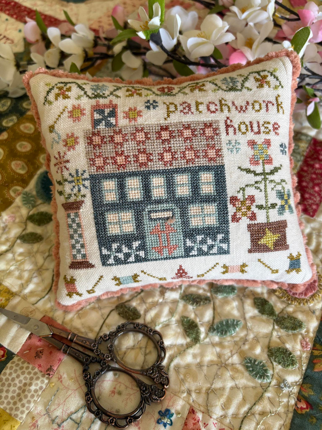 Decorative pillow with cross-stitch design featuring a house and flowers, placed on a quilt with scissors and flowers.