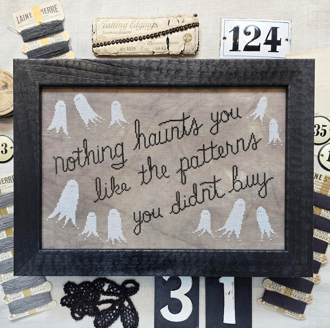 A framed cross-stitch piece with a black wood frame, featuring the phrase 'nothing haunts you like the patterns you didn't buy' in black script on a beige fabric background, surrounded by white ghost motifs. The frame is displayed on a styled craft table with spools of thread, a ribbon, and decorative numbers.