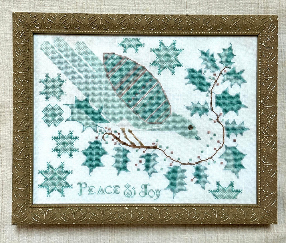 Framed cross-stitch artwork featuring a bird with a striped wing, surrounded by teal snowflakes, holly leaves, and a brown branch with red berries. The words 'PEACE & JOY' are stitched at the bottom.