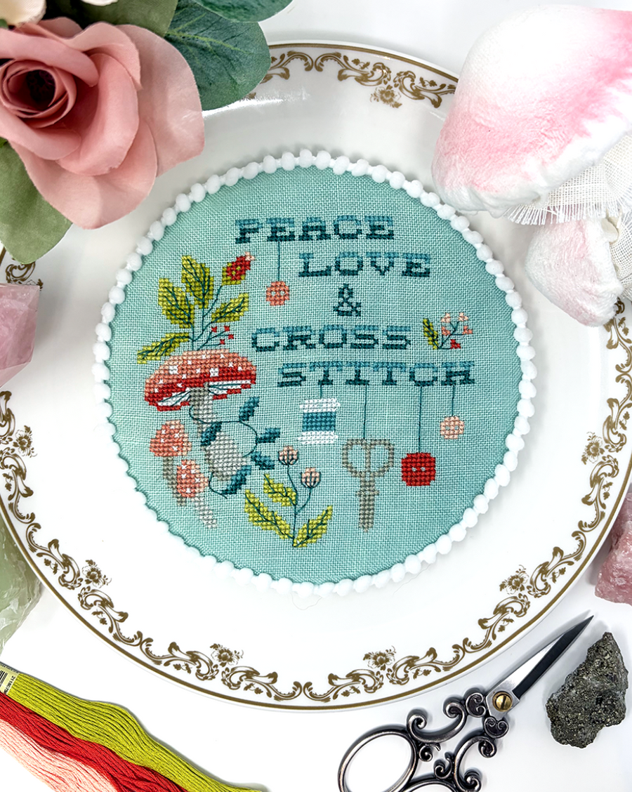 A round cross-stitch sampler with the words 'PEACE LOVE & CROSS STITCH' and illustrated mushrooms, flowers, and keys on a light blue background, displayed on an ornate white plate surrounded by pink roses, crystals, scissors, and thread.