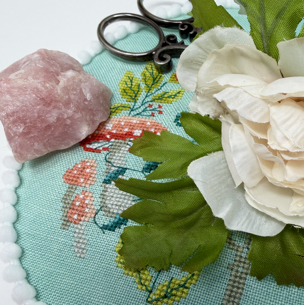 Cross-stitched fabric with flowers, scissors, and a pink stone on a white background.