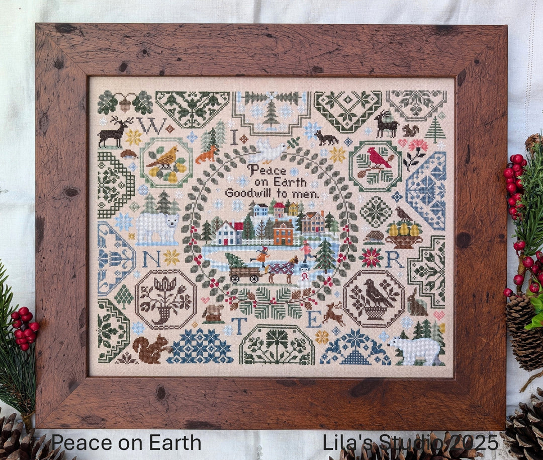 Framed cross-stitched sampler with 'Peace on Earth' text and festive elements, Lila's Studio branding visible.