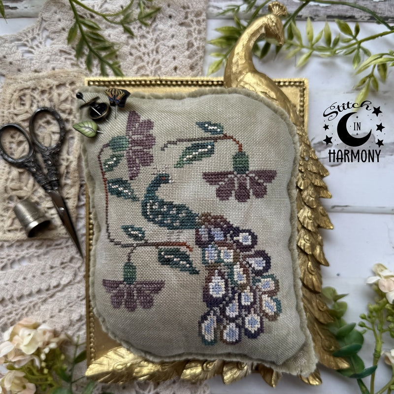 A square cross-stitch cushion with a peacock and floral design, styled on a white wooden surface with lace, greenery, scissors, and a bell. A gold peacock-shaped frame surrounds the cushion, and a 'Sutter in Harmony' logo is visible on the right.